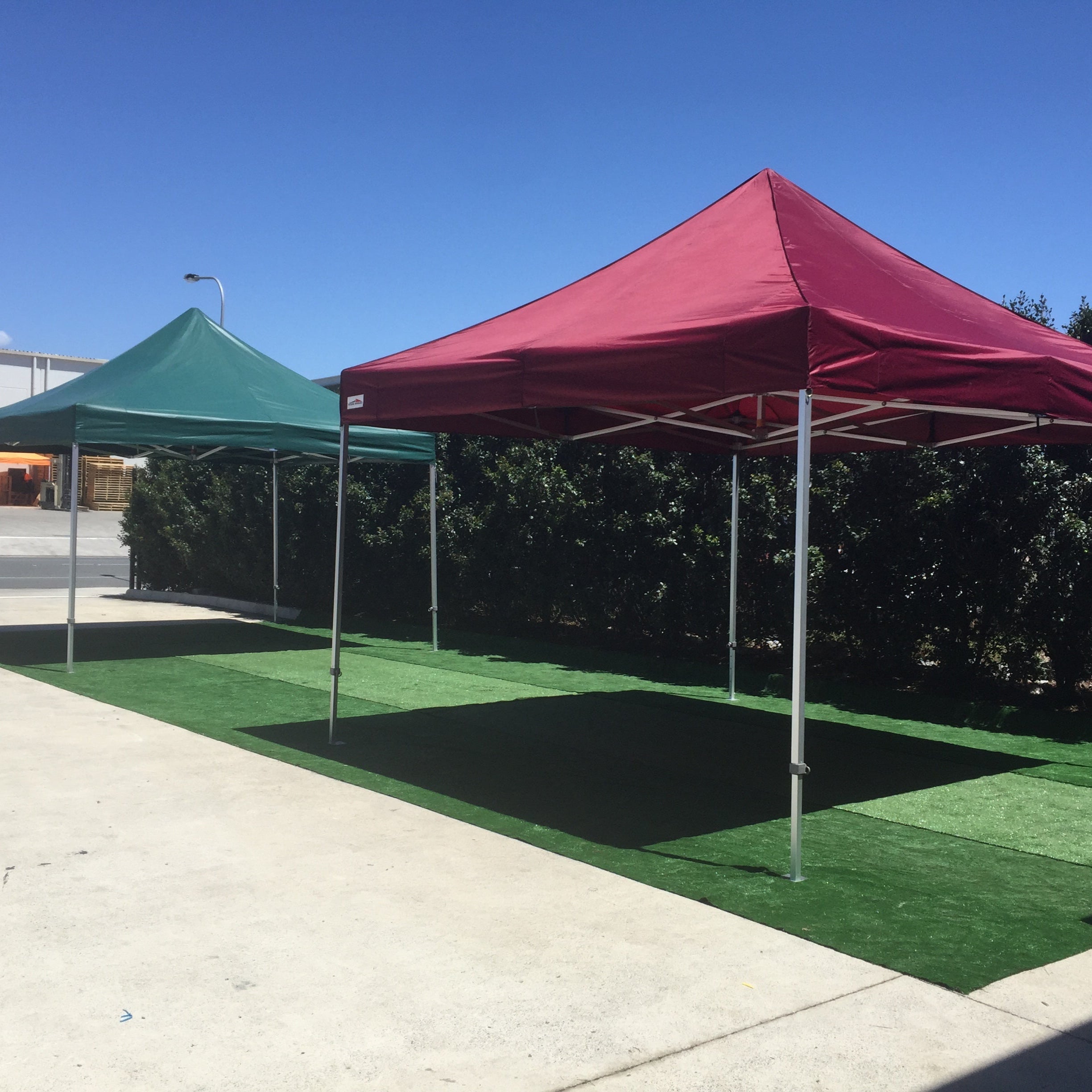 Green and maroon 4x4m X7 Tectonic canopy tents set up outdoors on artificial grass under a clear blue sky.