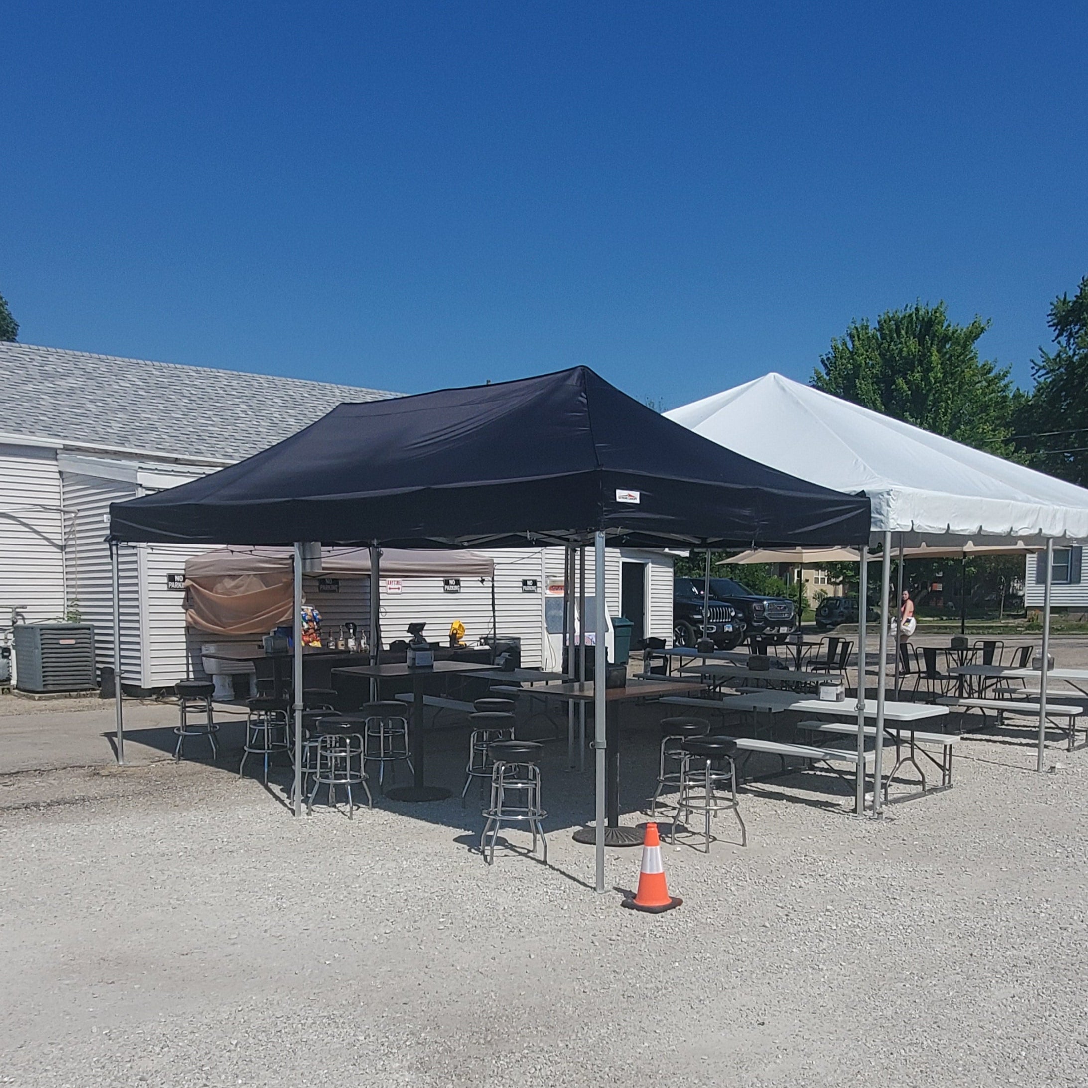 Black and white canopy tents set up outdoors on a gravel lot, providing shaded seating with tables and stools under clear blue skies.